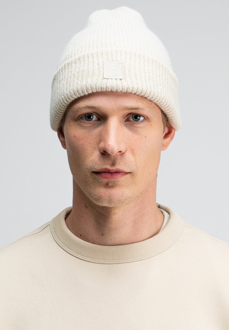 Weißer gerippter Strickbeanie mit einem kleinen Logo-Patch, kombiniert mit einem beigen Rundhals-Sweatshirt. Einfaches Design, weicher Stoff.