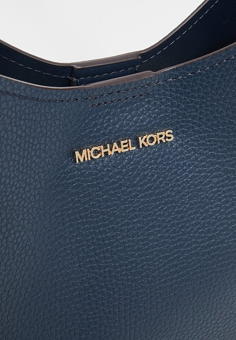 Gros plan d'un sac à main en cuir texturé bleu marine avec le logo doré "Michael Kors" et des coutures visibles le long du bord supérieur.