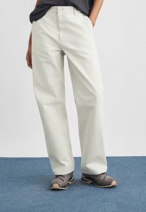 Persoană îmbrăcată cu pantaloni largi din catifea cu reavăn de culoare crem, bluză neagră și pantofi de drumeție gri, stând pe o mochetă albastră.