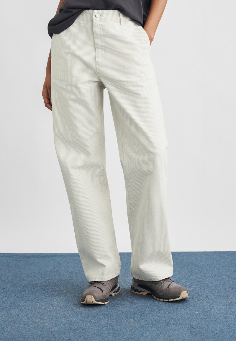 Person iført fløjlsbukser med brede ben i cremefarve, sort top og grå vandresko, stående på blåt tæppe.