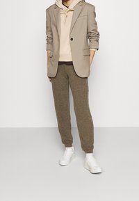 Beige blejzr přes béžovou mikinu s kapucí, kombinovaný s hnědými texturovanými tepláky a bílými teniskami. Blejzr má jeden knoflík a dvě kapsy.