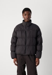 Svart boblejakke med høy krage, glidelås og horisontal quiltedesign. Har elastiske mansjetter og en liten logo.