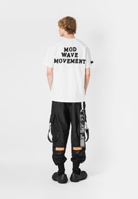 Camiseta blanca con el texto negro "MOD WAVE MOVEMENT" en la espalda; combinada con pantalones cargo negros y zapatos negros sin cordones.