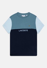 Tričko s barevným blokováním v jemně modré, tyrkysové a námořnické modři. Obsahuje zelené vyšívané logo a slovo "LACOSTE" v bílé barvě. Bavlněný materiál.