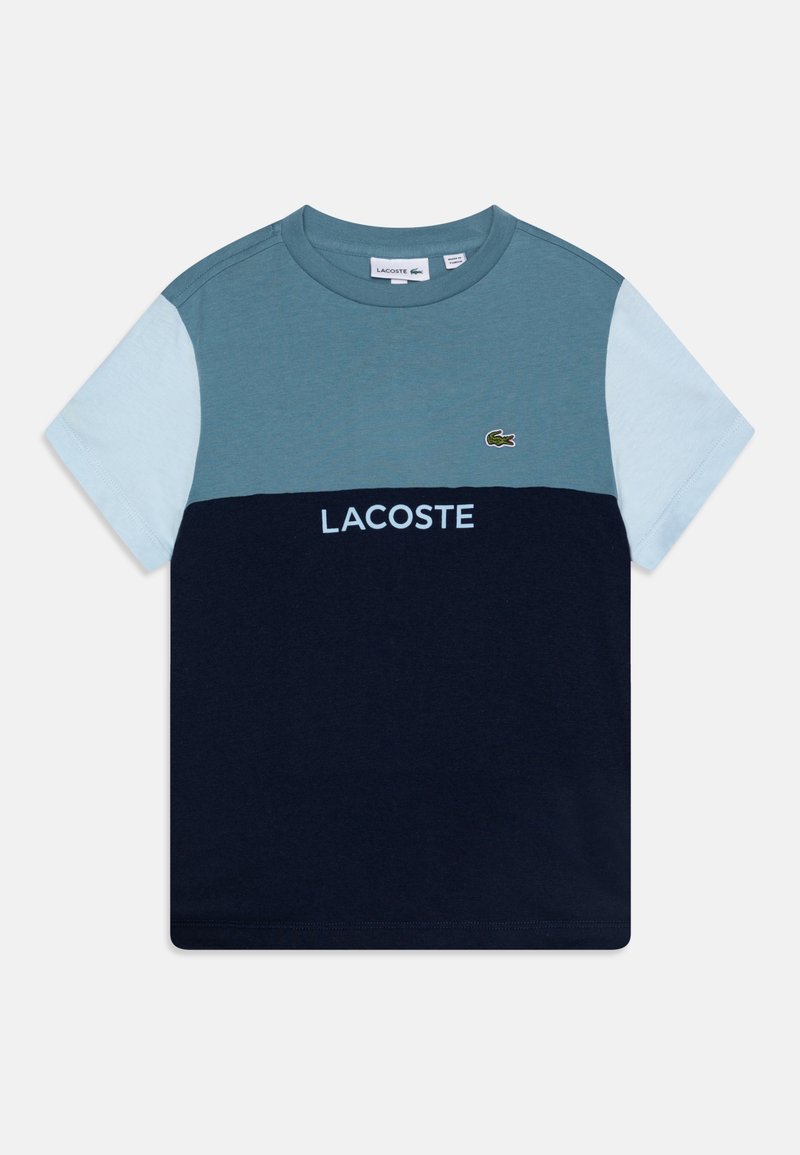 Tričko s barevným blokováním v jemně modré, tyrkysové a námořnické modři. Obsahuje zelené vyšívané logo a slovo "LACOSTE" v bílé barvě. Bavlněný materiál.