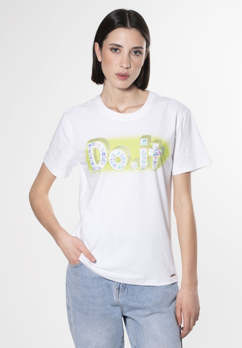 Camiseta blanca de algodón con un audaz texto "Hazlo" en letras multicolores con purpurina, sobre un fondo verde claro.
