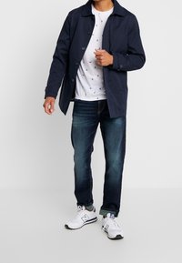 Marinblå regnjacka med krage, knappstängning, bärs över en vit t-shirt med små blå mönster, kombinerad med mörk denimjeans och vita sneakers.