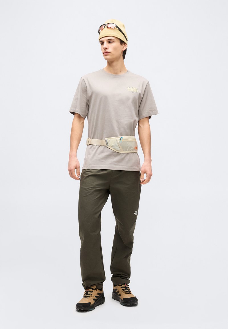 Young man wearing beige t-shirt, olive pants, tan hiking shoes, beige bucket hat with sunglasses, and a beige waist bag, standing against plain background.