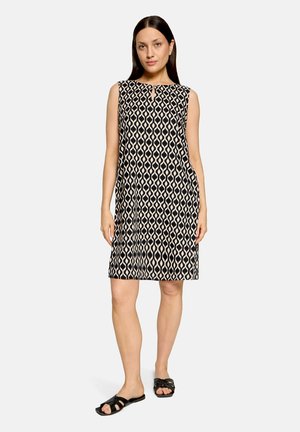 Femme debout portant une robe sans manches mi-longue noire et beige à motifs géométriques avec des sandales noires sans lacets sur fond blanc.