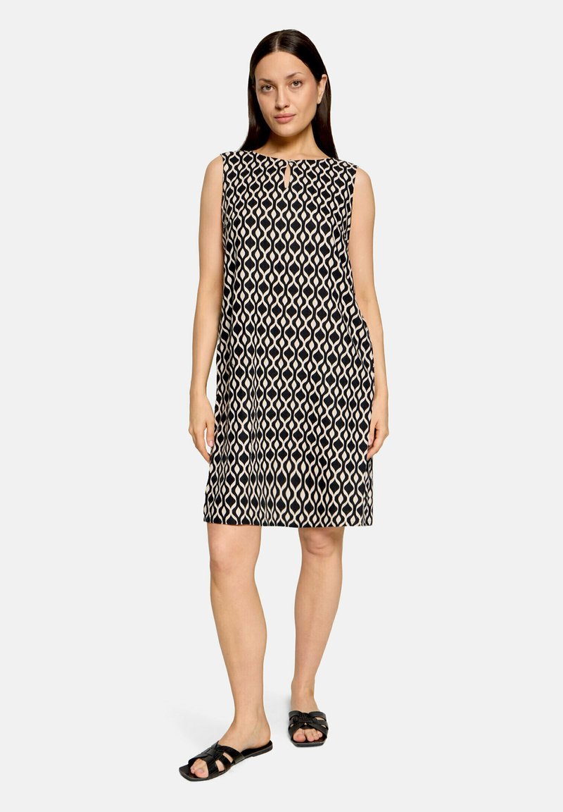 Femme debout portant une robe sans manches mi-longue noire et beige à motifs géométriques avec des sandales noires sans lacets sur fond blanc.