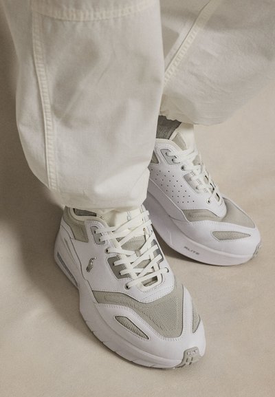 Λευκά και γκρί αθλητικά παπούτσια με κορδόνια φορεμένα με λευκό παντελόνι, εμφανίζοντας λεπτομέρειες των παπουτσιών όπως πάνελ από δίχτυ και λογότυπα της μάρκας σε ουδέτερο πάτωμα.