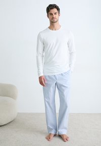 Tricou alb cu mânecă lungă asortat cu pantaloni de pijama rayati, de un albastru deschis, dintr-un amestec de bumbac moale. Stând pe un fundal de covor bej.