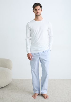 Tricou alb cu mânecă lungă asortat cu pantaloni de pijama rayati, de un albastru deschis, dintr-un amestec de bumbac moale. Stând pe un fundal de covor bej.