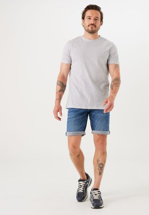 T-shirt gris à manches courtes avec motif de vagues, shorts en denim retroussés au niveau des ourlets, et baskets marines avec lacets blancs, portés par un homme tatoué.