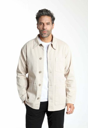 Veste en toile beige clair avec fermeture à boutons, poche poitrine, deux poches devant et col structuré. Portée par-dessus une chemise blanche.