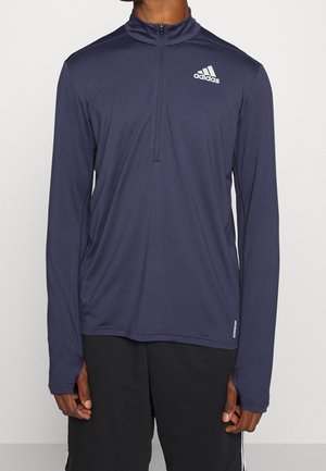 Man wearing navy long-sleeve half-zip Adidas sports top and black Adidas shorts, standing against a plain light background.