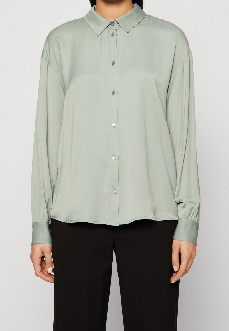Femme portant une chemise boutonnée en satin vert clair, ample et à manches longues, et un pantalon noir à jambe droite, debout devant un fond uni.