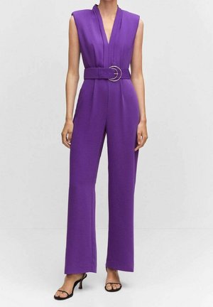 Hihaton violetti haalari, jossa syvä V-pääntie, leveä vyö kaksoissoljella, leveälahkeiset housut, yhdistetty mustiin avonaisiin korkokenkiin.