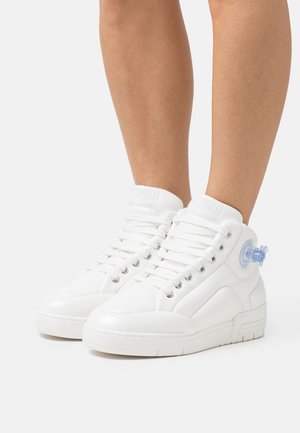 Personne portant des baskets montantes blanches avec des lacets blancs et un petit détail bleu translucide sur le côté extérieur de chaque chaussure.