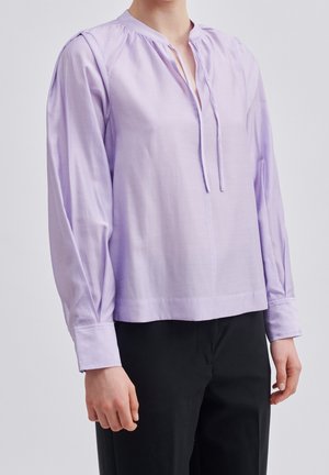 Persoon die een lichtpaarse blouse met lange mouwen en een gestrikte halslijn draagt, gecombineerd met een zwarte broek, tegen een effen achtergrond.