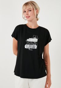 Sort kortærmet t-shirt med en løs pasform, prydet med et blomstermotiv i hvid og grå, samt ordet "Floraison" i bunden.
