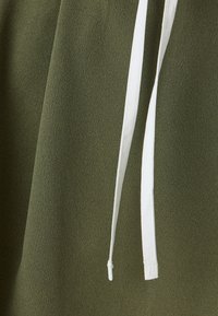 Tissu vert olive avec une texture lisse. Comprend un détail de cordon blanc pour l'ajustement. La vue rapprochée met en avant le matériau et le design.