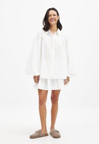 Balta lina oversized krekls ar pūkainām piedurknēm, ar pogām priekšā, apvienots ar baltiem slāņveida svārkiem un gaiši brūnām uzvelkamām kurpēm.