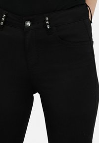 Fekete, rugalmas farmernadrág slim-fit szabással. Elülső zsebein fényes díszítésekkel és klasszikus gombos záródással rendelkezik.