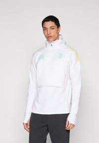 Fehér sportdzseki félcipzárral és kapucnival, menta zöld és sárga díszítésekkel, rózsaszín varrással és hüvelykujjnak kialakított lyukakkal az ujjakon.