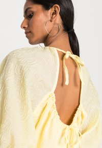 Top amarelo texturizado com um corte solto, costas abertas e fecho com laço. Detalhes notáveis incluem um padrão floral e mangas largas.