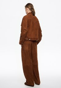 Giacca in suede marrone con yoke in stile western e pantaloni a gamba larga coordinati, dotati di ampie tasche e una sottile texture.