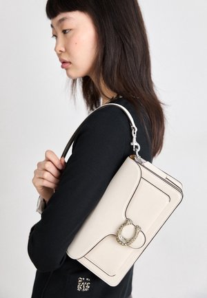Mujer con el pelo liso y oscuro, vestida de negro, sostiene un bolso de hombro color crema con cierre circular adornado con joyas, mirando hacia la izquierda sobre fondo claro.