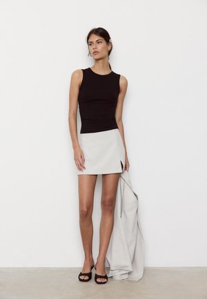 Kvinde iført sort ærmeløs top, hvid miniskørt med sideslids, sorte åbne hæle, holder en hvid blazer mod en ensfarvet hvid væg.