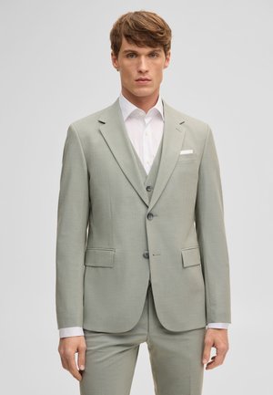 Giovane con capelli castani corti che indossa un completo tre pezzi grigio chiaro e una camicia bianca, su sfondo semplice.