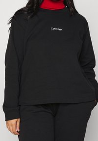 Svart sweatshirt i bomull med långa ärmar, rund halsringning och en liten vit "Calvin Klein" logotyp på bröstet. Slät textur.