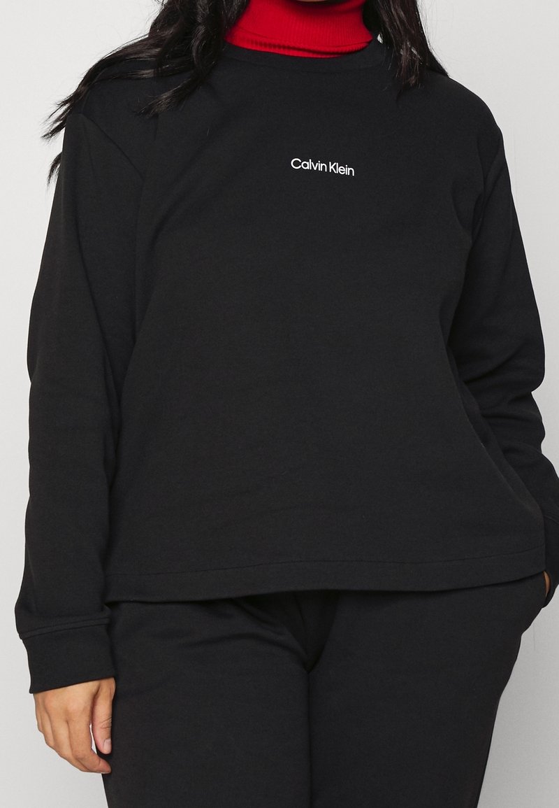 Svart sweatshirt i bomull med långa ärmar, rund halsringning och en liten vit "Calvin Klein" logotyp på bröstet. Slät textur.