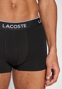 Sorte boksertruser laget av myk bomullsblanding, med hvit linning som har "LACOSTE"-logo. Tett passform, glatt tekstur.