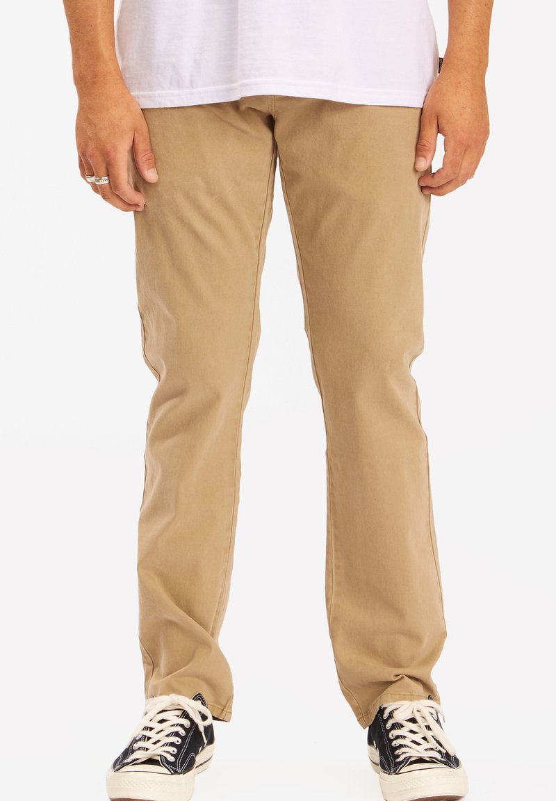 Chinos beige con un diseño de pierna recta, fabricados en algodón y con una textura sutil. Llevados con zapatillas negras que tienen cordones y acentos en blanco.