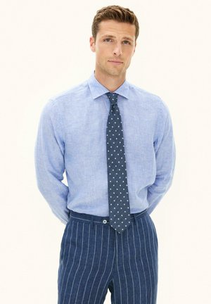 Mand iført lyseblå skjorte, navyblåt slips med polkaprikker og navyblå benklæder med striber, stående med hænderne på ryggen.