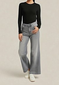 Femme portant un haut à manches longues noir, un jean gris taille haute à jambes larges et des baskets blanches, debout avec une main dans la poche.