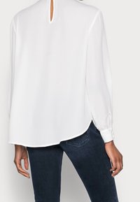 Blouse blanche à manches longues avec un col rond, une découpe en forme de goutte d'eau dans le dos et des poignets boutonnés. Le tissu semble lisse et léger. Associée à un jean foncé.