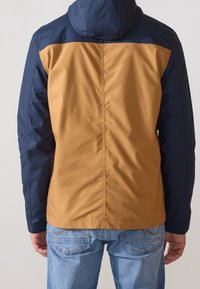 Färgblockerad jacka med marinblå och beige paneler, huva och en slät textur. Bärs med blå jeans och visar en tvåriktsdesign.