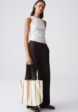 Frau trägt ärmelloses weißes Oberteil und weite schwarze Hose und hält eine große beige Tragetasche mit schwarzen vertikalen Streifen.