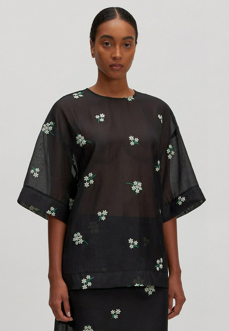 Schwarze durchsichtige Bluse mit kurzen, weiten Ärmeln, die mit weißen Blumenstickereien und grünen Stielen verziert ist. Glatte Textur, lässiges Design, leichtes Material.
