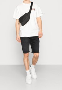Riñonera negra con cremallera, llevada sobre una camiseta blanca con un logo multicolor, combinada con pantalones cortos de mezclilla negros y zapatillas blancas.