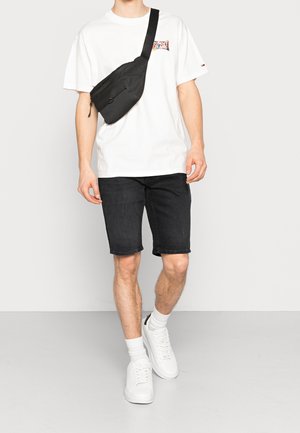 Riñonera negra con cremallera, llevada sobre una camiseta blanca con un logo multicolor, combinada con pantalones cortos de mezclilla negros y zapatillas blancas.