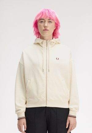 Person mit leuchtend pinken Haaren, die einen cremefarbenen Kapuzenpullover mit Reißverschluss und kleinem gesticktem Logo sowie schwarze Hosen trägt, steht vor einem einfarbigen Hintergrund.