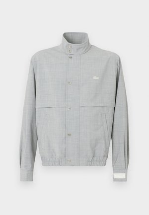 Veste grise clair en tissu respirant, dotée d'un col montant, d'une fermeture à boutons-pression et de poignets élastiques ; comporte un petit logo.