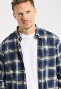 Homme aux cheveux blonds courts portant une chemise à carreaux bleus et blancs par-dessus un tee-shirt blanc uni, regardant vers la caméra.