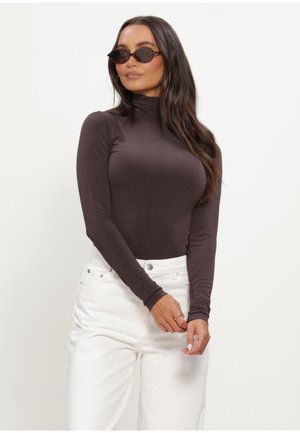 Vrouw met lang donker haar die ovale zonnebrillen draagt, een aansluitende donkerbruine coltrui, witte hoge taillebroeken en ringen aan haar vingers.